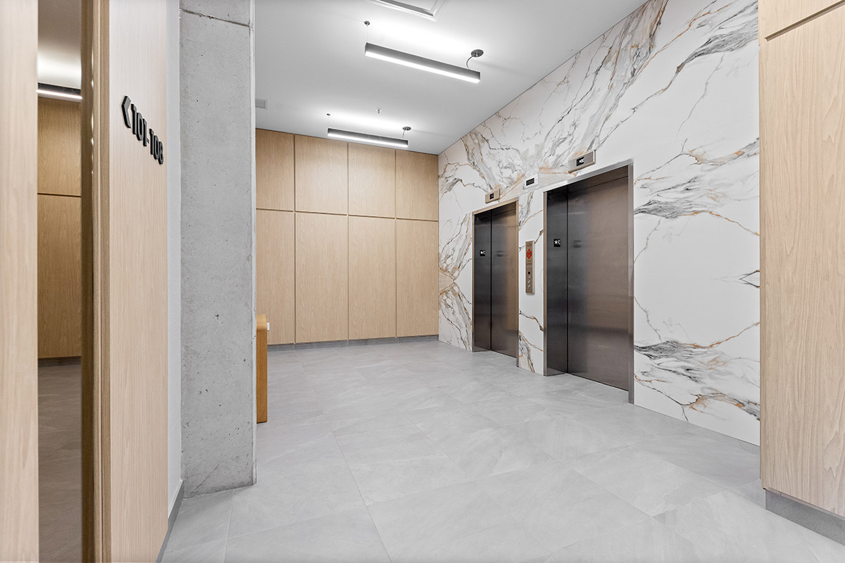 Hall d’ascenseurs moderne avec finitions en marbre et panneaux de bois pâle, comprenant deux ascenseurs.